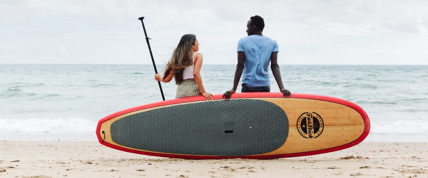 Two students at the beach talking and holding together a stand up paddle board