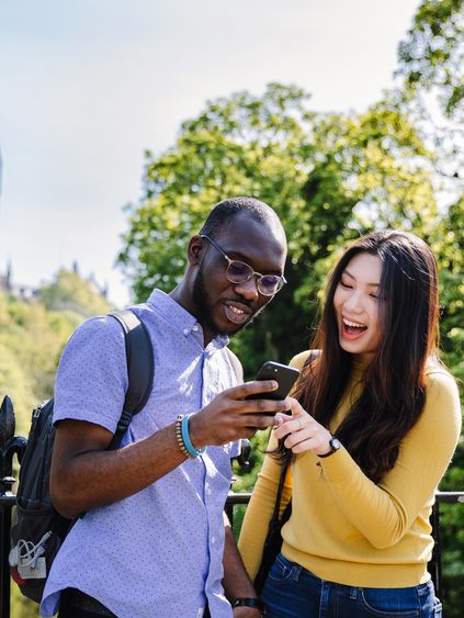 Students looking at a phone with Gilbert Scott building behind together with trees and greenery