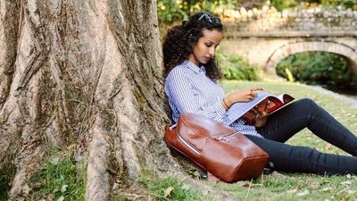 A woman sits against a tree by a river reading