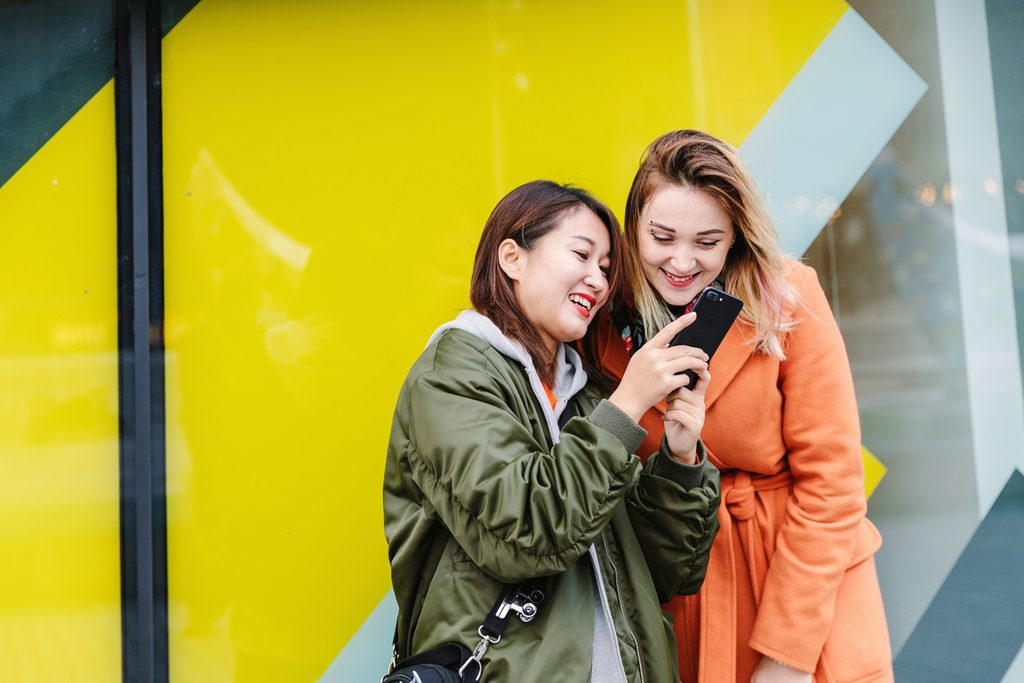 Two international students, captivated by their phones, in front of a yellow building.