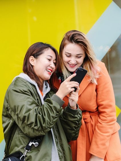 Two international students, captivated by their phones, in front of a yellow building.