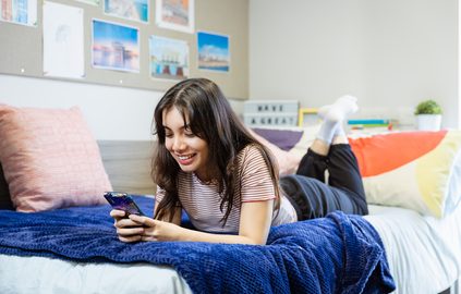 A picture of a student relaxing on her bed, using her phone to text.
