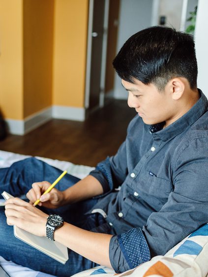 A student watching a lecture recording, taking notes on a bed.