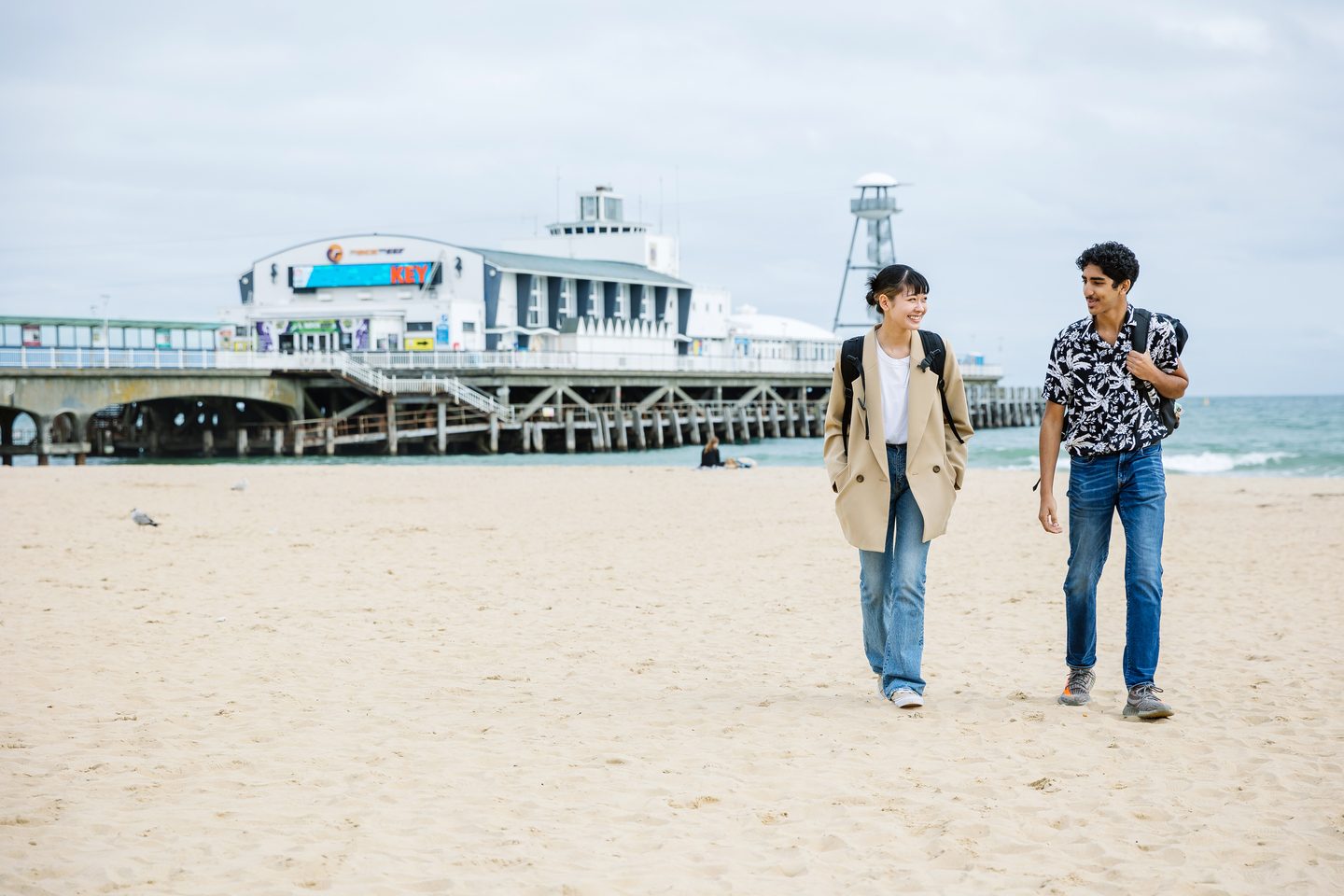 A male student and a female student smile as they walk along a sandy Bournemouth beach, with Bournemouth Pier behind them.